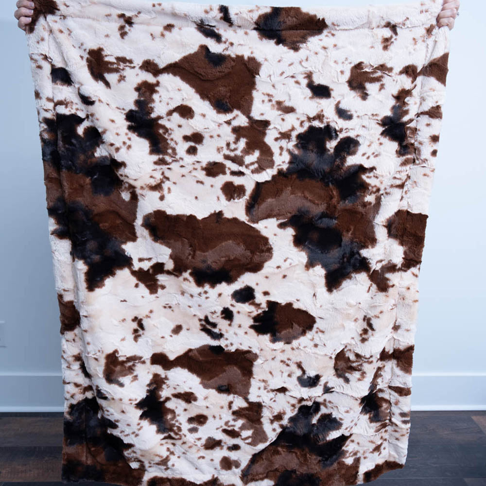 Brown and white cow print blanket held by a person against a white wall.
