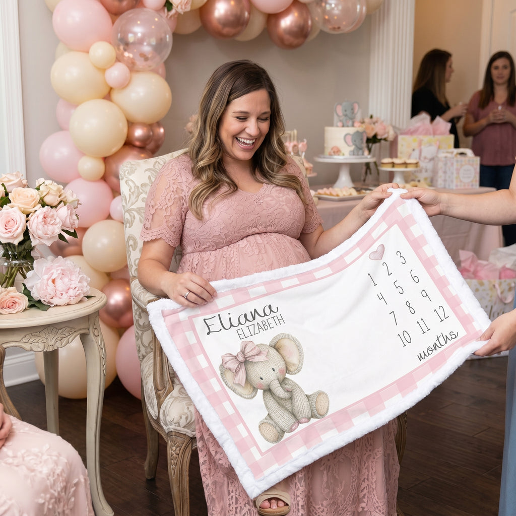 A pregnant woman in a pink dress holds a personalized baby blanket with a pink gingham border, a gray elephant illustration, and the name 'Eliana Elizabeth' along with dates and location text. The scene is set in a decorated baby shower or birthday party with pink and gold balloons, flowers, and cake tables in the background.