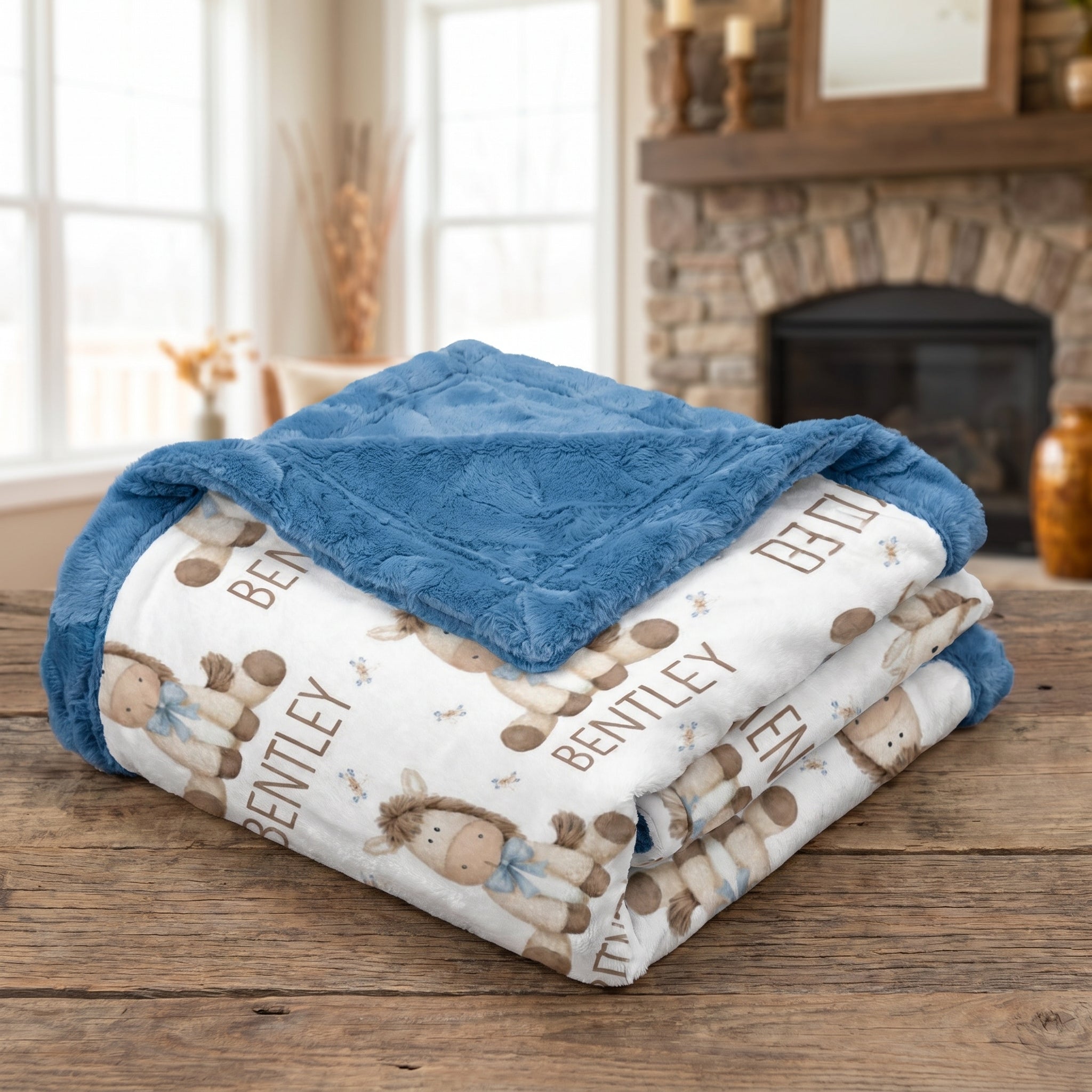 A folded baby blanket with a blue plush reverse side and a white front side printed with teddy bears and the name 'BENTLEY', displayed on a wooden table in front of a cozy fireplace.