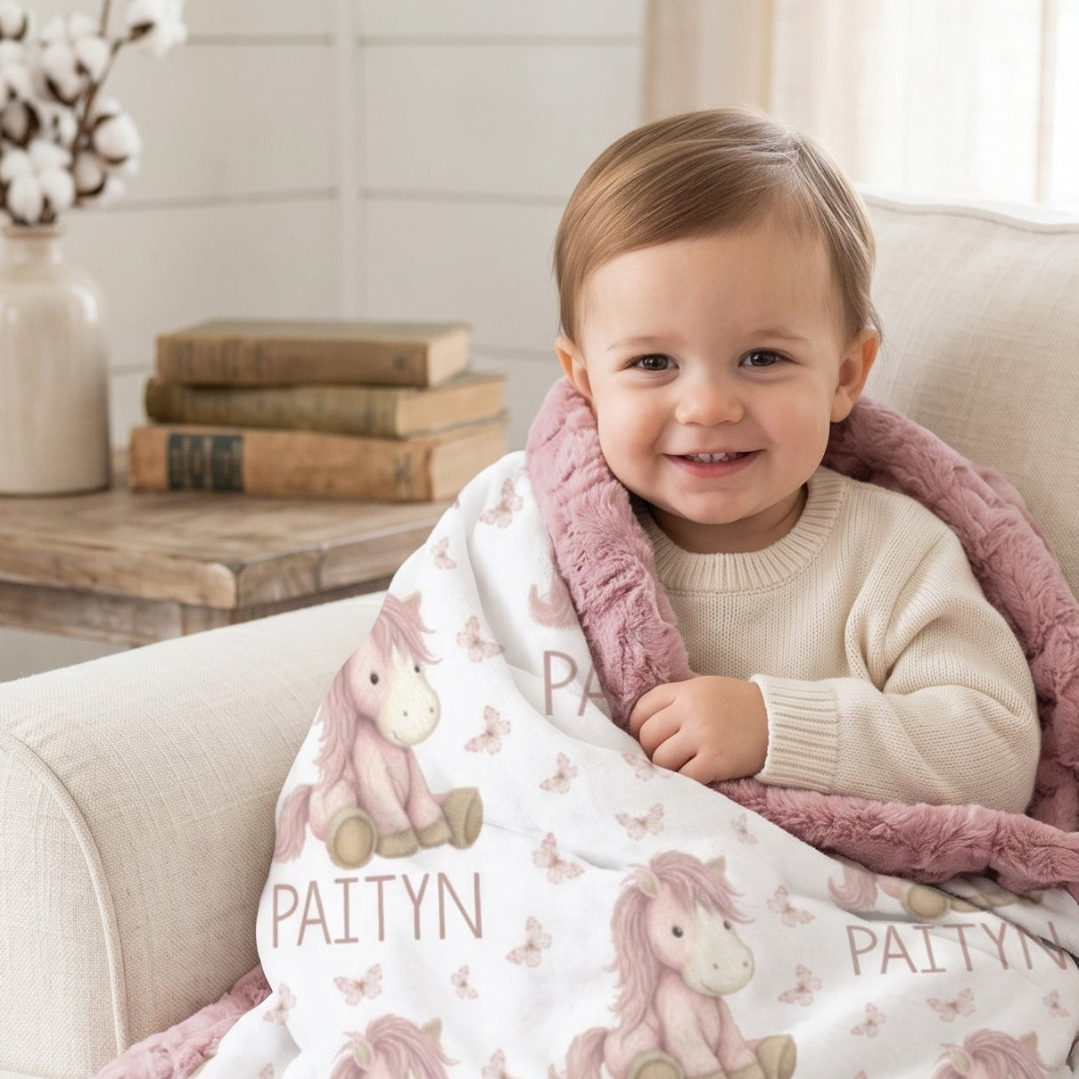 A baby sitting on a cream sofa wrapped in a personalized blanket. The blanket has a white front with a pink unicorn pattern and the name 'PAITYN' printed repeatedly, finished with a soft pink plush border.