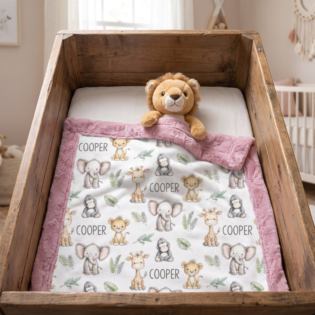 A personalized baby blanket with a white background, pink fuzzy border, and cute animal illustrations, styled inside a rustic wooden bassinet with a plush lion toy.
