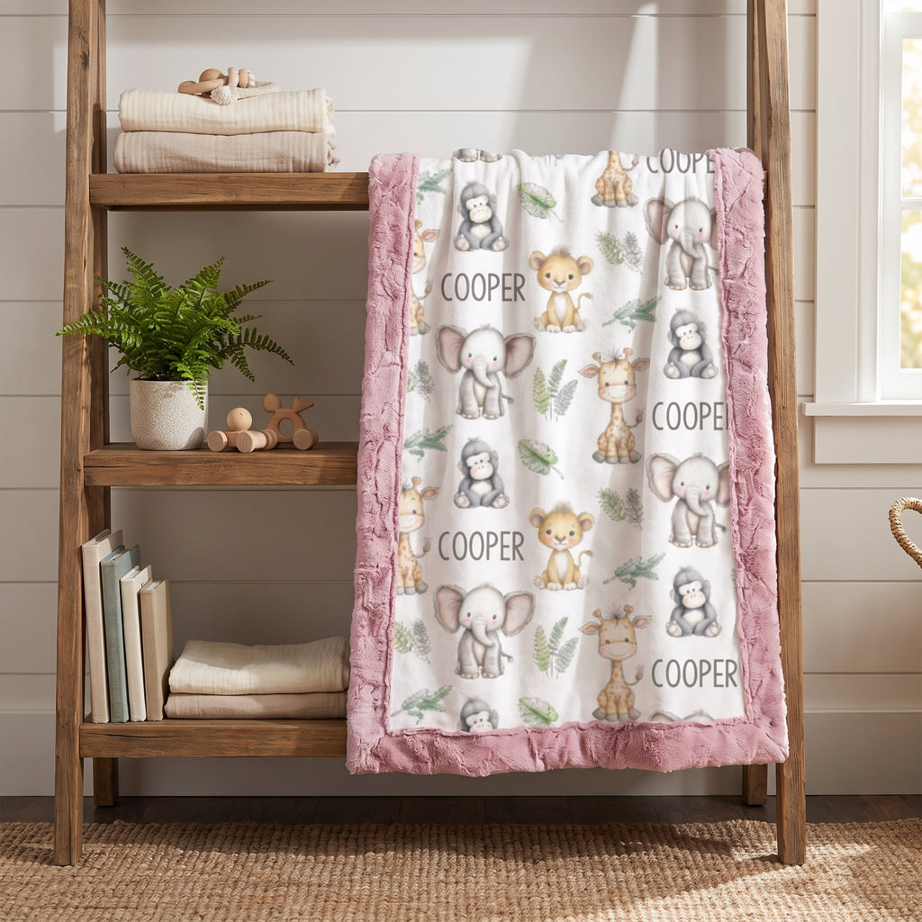 A baby blanket with a pink fringed border and an all-over print of cute cartoon animals and the name 'COOPER', draped over a wooden shelf in a nursery setting.