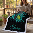 A woman sits on a wooden porch swing reading a book, with a black fleece throw blanket draped over the bench. The blanket features a turquoise and lime green paddle-and-ball graphic and the name 'Trena' in script.