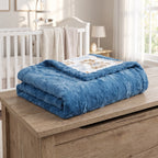 A folded blue plush blanket with a teddy bear illustration on the corner is displayed on a wooden chest in a softly lit nursery setting.