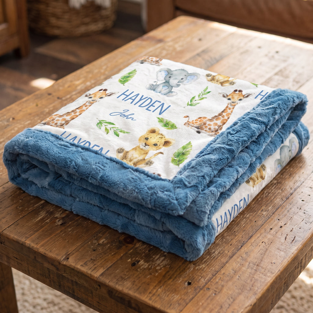 A personalized baby blanket with a blue plush border and a white printed center featuring safari animals like giraffes, a lion, and an elephant, folded on a rustic wooden table. The blanket is personalized with the name 'HAYDEN' and the name 'John'.