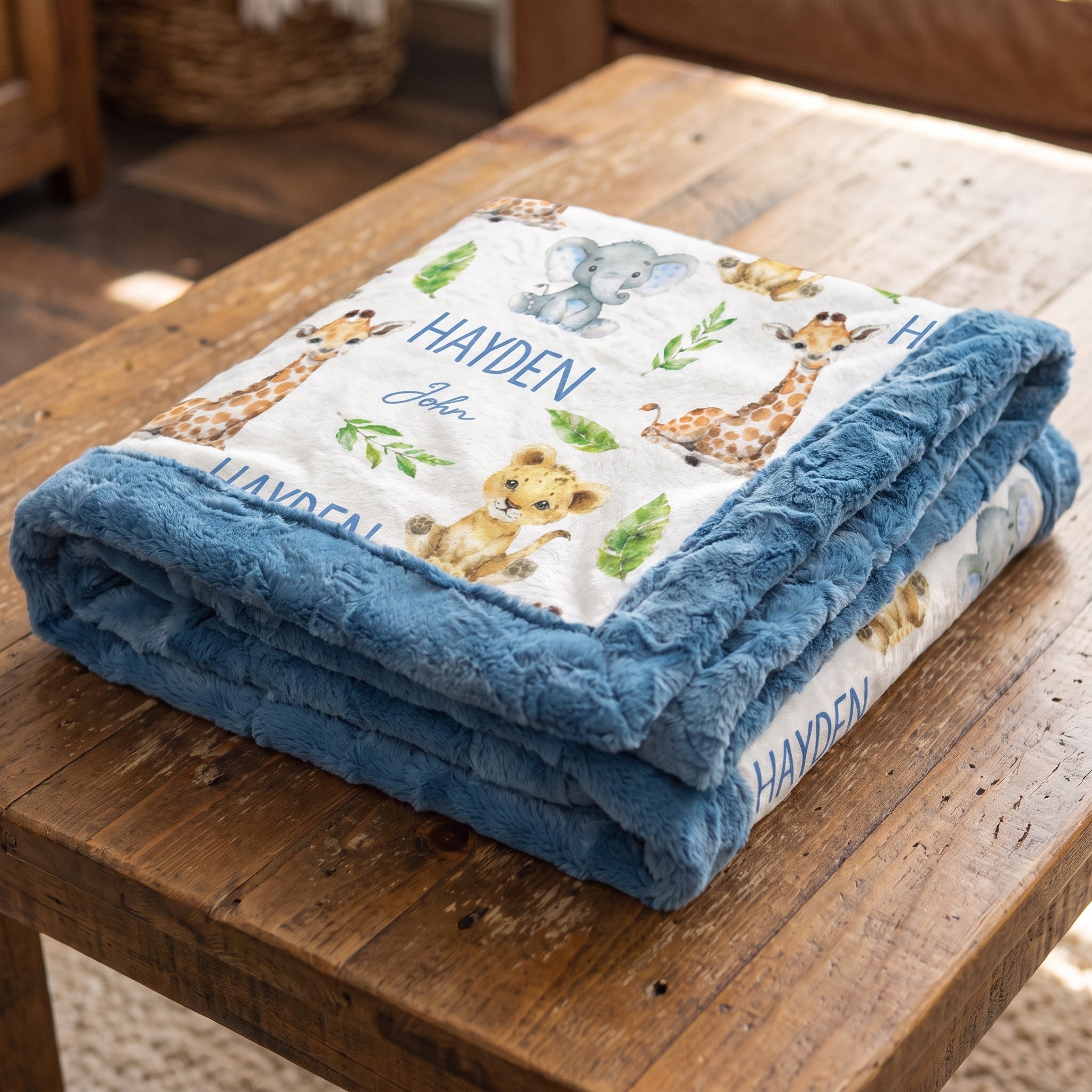 A personalized baby blanket with a blue plush border and a white printed center featuring safari animals like giraffes, a lion, and an elephant, folded on a rustic wooden table. The blanket is personalized with the name 'HAYDEN' and the name 'John'.