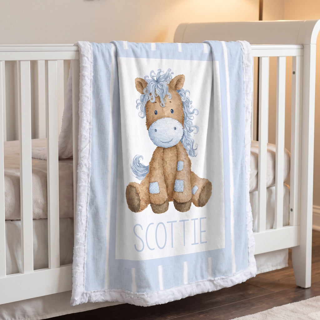 A light blue and white fringed baby blanket draped over a white crib, featuring a watercolor-style brown horse illustration with a blue mane and the name 'SCOTTIE' printed below.