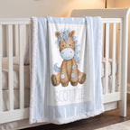 A light blue and white fringed baby blanket draped over a white crib, featuring a watercolor-style brown horse illustration with a blue mane and the name 'SCOTTIE' printed below.