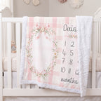 A personalized baby blanket draped over a white crib, featuring a pink and white gingham pattern, a floral wreath frame, and a printed baby name and monthly timeline.
