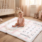 A baby sits on a personalized monthly calendar floor mat in a nursery. The mat features a pink and white gingham pattern, a floral wreath design, and a space to write the baby's name and count from 1 to 12 months.