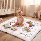 A baby sits on a personalized baby blanket in a bright nursery. The blanket features a pink and white gingham pattern, a white fuzzy border, and a printed number calendar from 1 to 12 with the name 'Quisey' written at the top right.