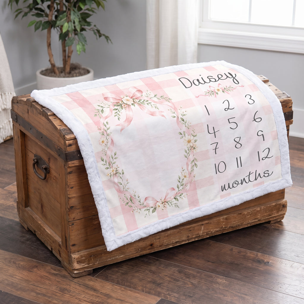 A personalized baby blanket with a pink and white gingham pattern, a floral wreath design, and the name 'Daisy' along with a numbered months chart, draped over a rustic wooden chest on a hardwood floor.