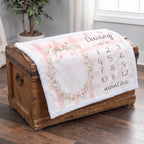 A personalized baby blanket with a pink and white gingham pattern, a floral wreath design, and the name 'Daisy' along with a numbered months chart, draped over a rustic wooden chest on a hardwood floor.