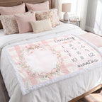 A personalized baby blanket with a pink and white gingham pattern, a floral wreath design, and the name 'Daisy' along with months 1 through 12, styled on a bed with matching pillows.