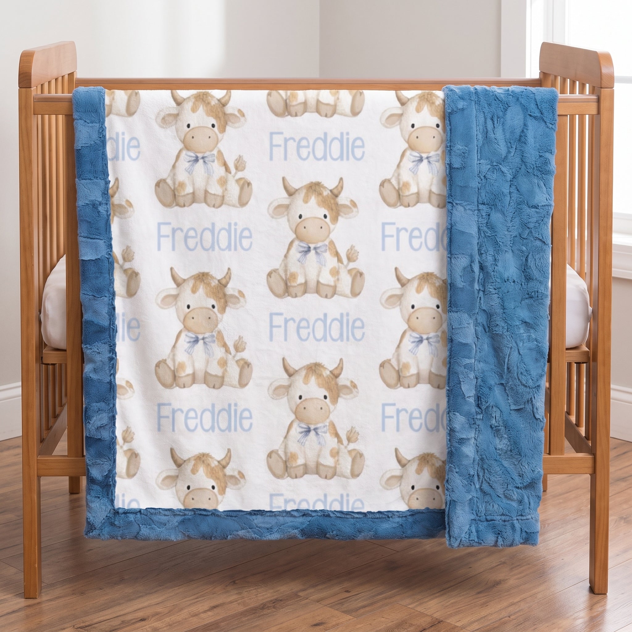 A baby crib bedding set featuring a white quilted top sheet with a repeating pattern of cute light brown and white cows wearing blue bows, the name 'Freddie' printed in light blue, and a plush blue ruffled bumper. The crib is made of natural wood and is set in a bright room with a wooden floor.