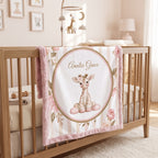A personalized baby blanket draped over a wooden crib in a softly lit nursery. The blanket features a central oval design with a cute cartoon giraffe and the names 'Amelia Grace' in script, surrounded by pink flowers and green leaves.
