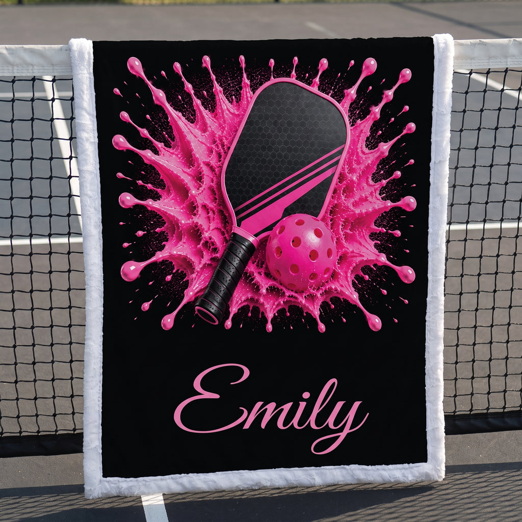 A personalized black fleece blanket with a white fringed border, featuring a pink and black pickleball graphic with a pink paint splatter background and the name 'Emily' in pink script. The blanket is draped over a tennis net on an outdoor court.