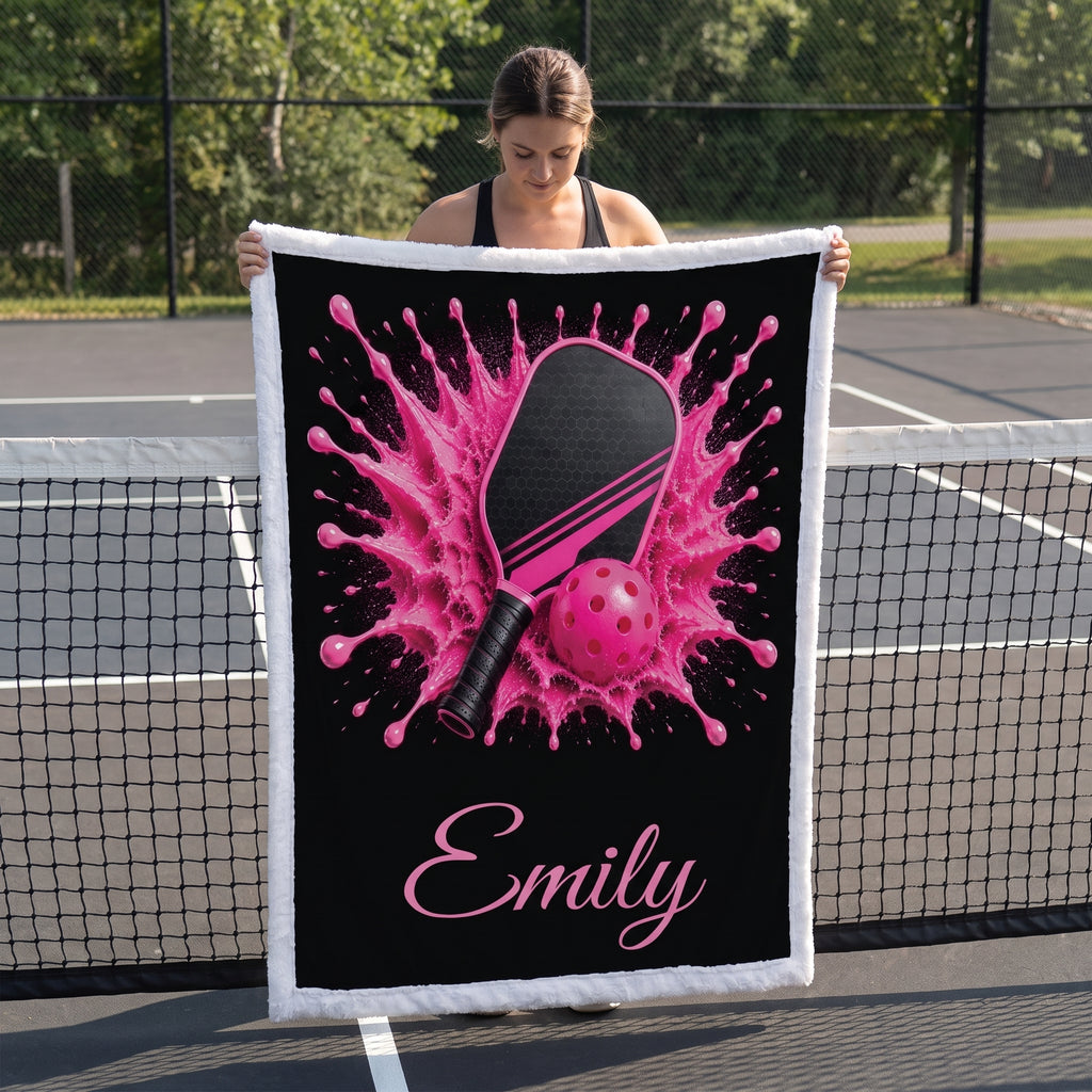 A woman stands on a tennis court holding up a custom-printed throw blanket. The blanket features a black background with a vibrant pink table tennis splash design, a pink paddle, and a pink ball, with the name 'Emily' printed in pink script at the bottom.