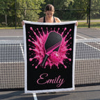 A woman stands on a tennis court holding up a custom-printed throw blanket. The blanket features a black background with a vibrant pink table tennis splash design, a pink paddle, and a pink ball, with the name 'Emily' printed in pink script at the bottom.