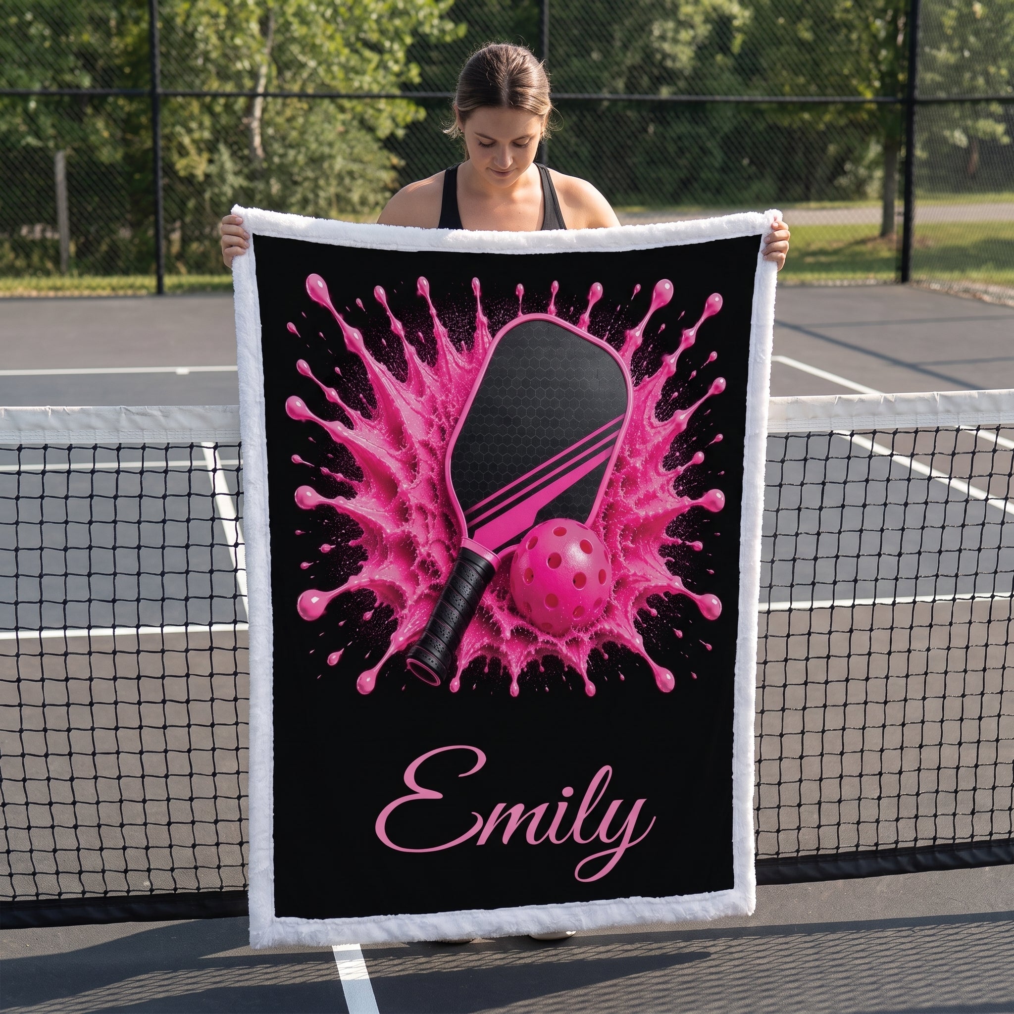 A woman stands on a tennis court holding up a custom-printed throw blanket. The blanket features a black background with a vibrant pink table tennis splash design, a pink paddle, and a pink ball, with the name 'Emily' printed in pink script at the bottom.