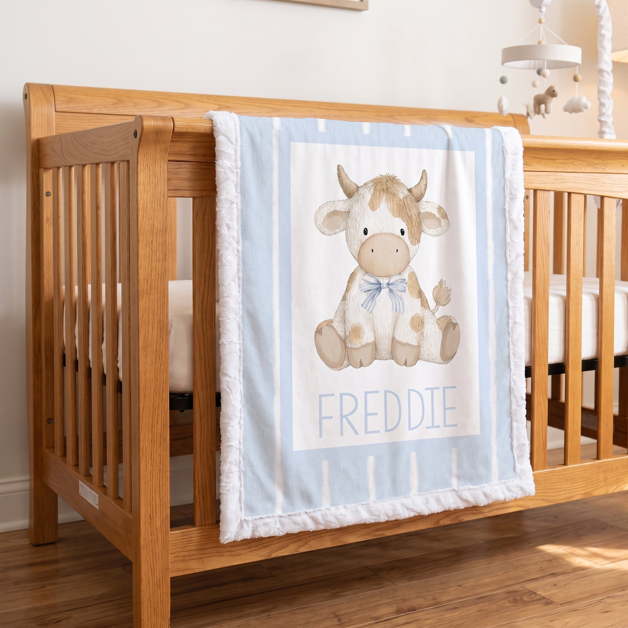 A personalized baby blanket draped over a wooden crib, featuring a watercolor-style cow illustration and the name 'FREDDIE' in blue text, set in a nursery room.
