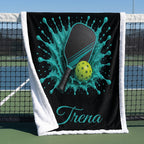 A personalized black fleece blanket with a white sherpa border, featuring a custom graphic of a pickleball paddle and ball with a turquoise splash design and the name 'Trena' in script. The blanket is draped over a tennis net on an outdoor court.