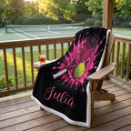 A personalized black fleece blanket with a pink tennis splash design, a tennis racket graphic, a tennis ball, and the name 'Julia' in pink script, draped over a wooden lounge chair on a wooden deck with a tennis court in the background.