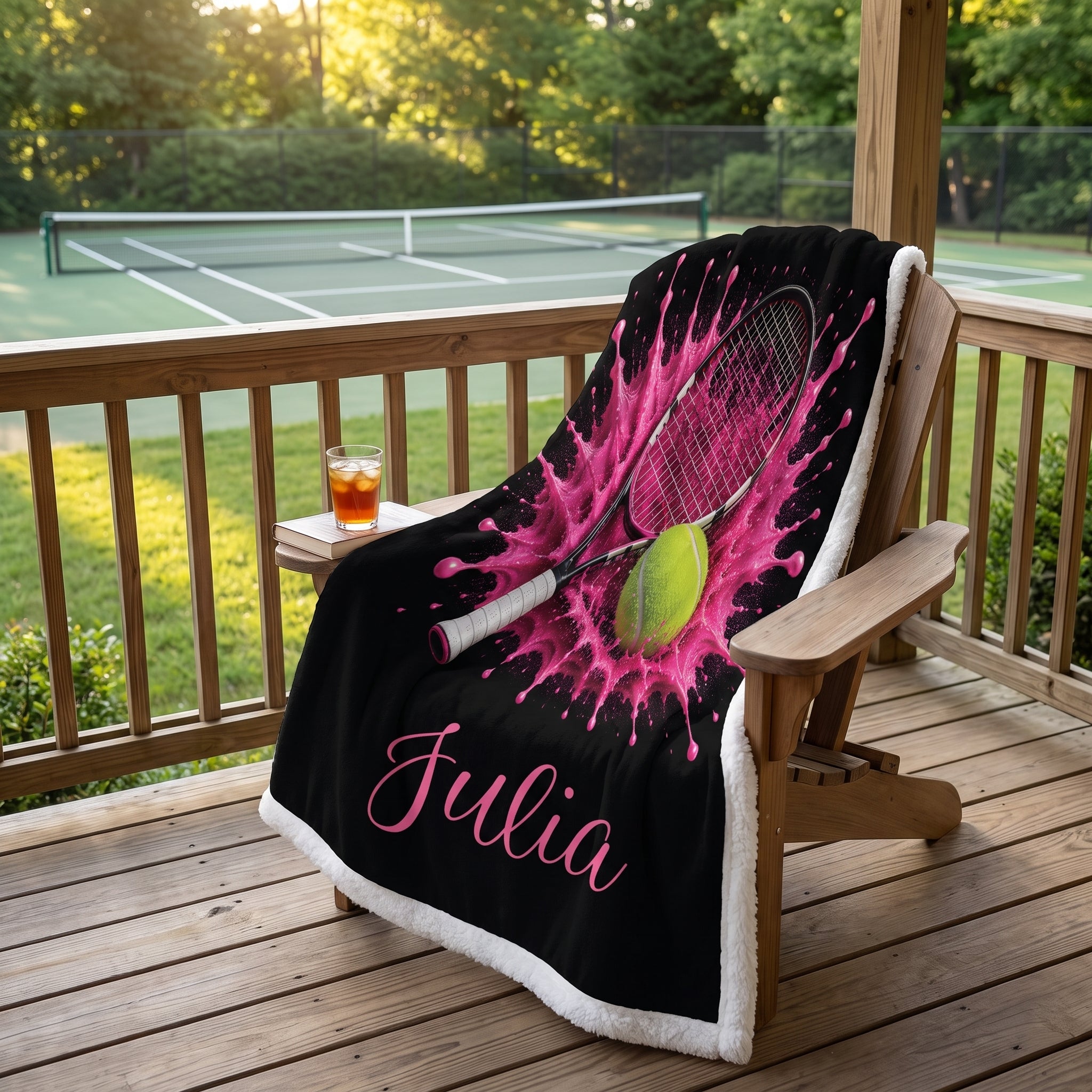 A personalized black fleece blanket with a pink tennis splash design, a tennis racket graphic, a tennis ball, and the name 'Julia' in pink script, draped over a wooden lounge chair on a wooden deck with a tennis court in the background.