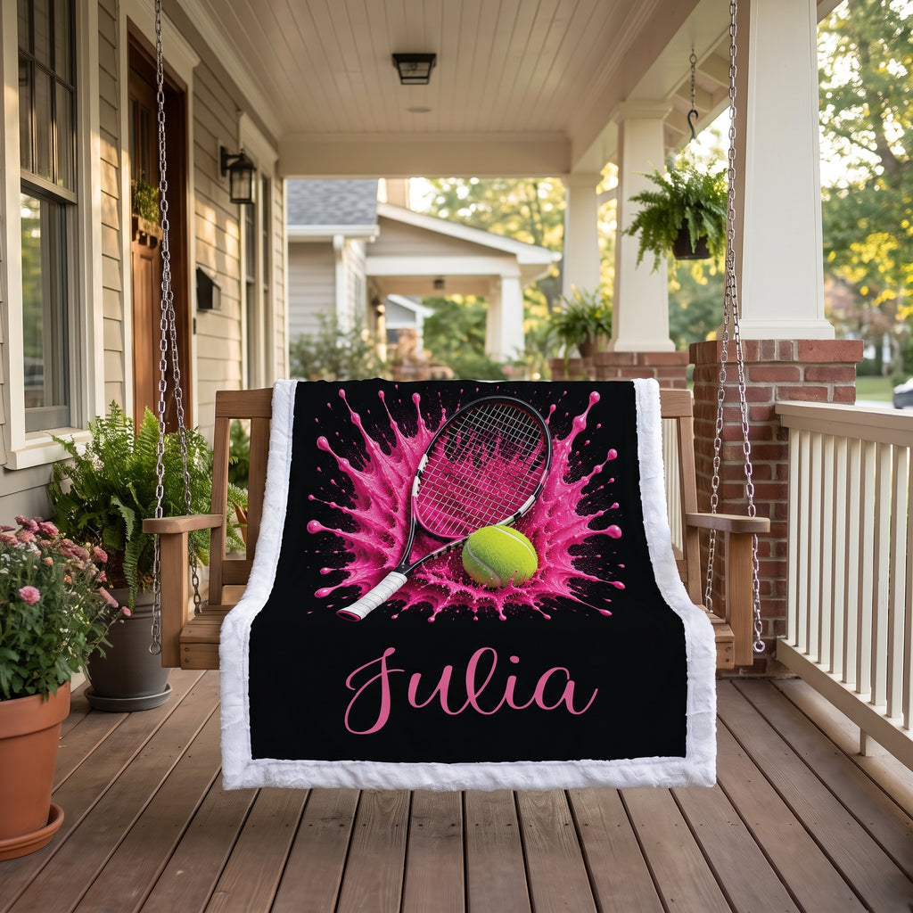 A custom black throw blanket with a white fringed border is draped over a wooden porch swing. The blanket features a pink tennis splash design, a tennis racket, a yellow tennis ball, and the name 'Julia' in pink script.
