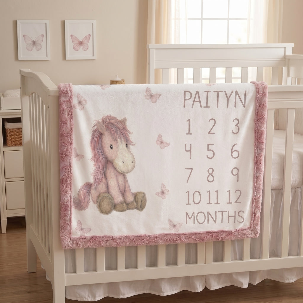 A personalized baby blanket draped over a white wooden crib in a nursery. The blanket is white with a pink fuzzy border, featuring a pink and brown unicorn illustration on the left and the name 'PAITYN' above a 12-month countdown from 1 to 12 on the right.