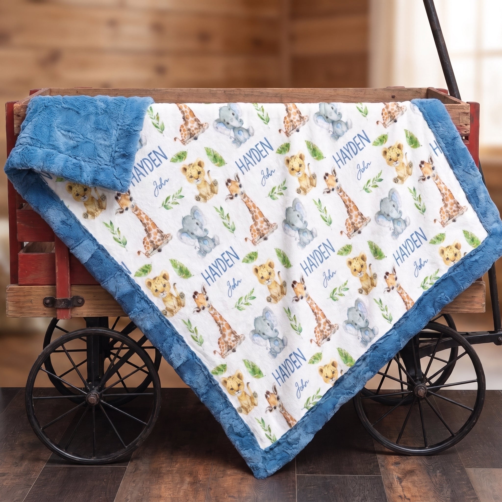 A plush baby blanket with a blue border and an all-over safari animal print featuring giraffes, lions, elephants, and the name 'Harden John' in blue script, draped over a rustic red wagon.