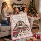 A woman and a girl hold up a personalized Christmas movie watching blanket in a cozy living room decorated for Christmas. The blanket features a pink buffalo plaid border, a central illustration of a rustic cabin with dogs and a red truck, and the text 'Personalized Christmas Movie Watching Blanket'.