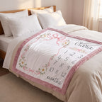 A personalized baby milestone blanket draped over a bed. The white blanket features a pink floral border, pink ribbon bows, and a printed black timeline of months from 1 to 12 along with the name 'Clara' in script.