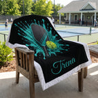 A custom black fleece blanket with a white sherpa border draped over a wooden chair on a patio. The blanket features a graphic of a ping-pong paddle and ball inside a teal paint splash, with the name 'Trena' written in teal script.