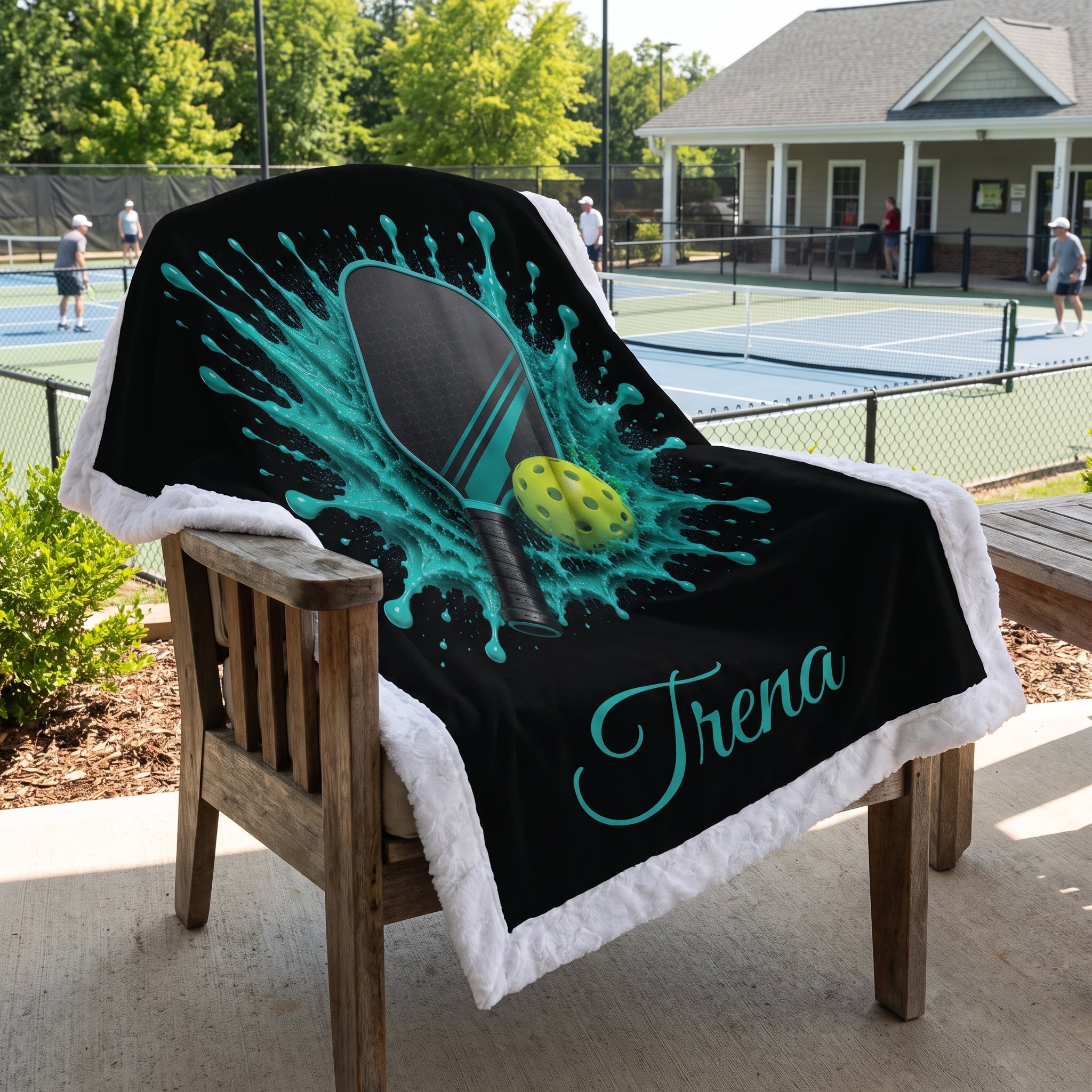 A custom black fleece blanket with a white sherpa border draped over a wooden chair on a patio. The blanket features a graphic of a ping-pong paddle and ball inside a teal paint splash, with the name 'Trena' written in teal script.
