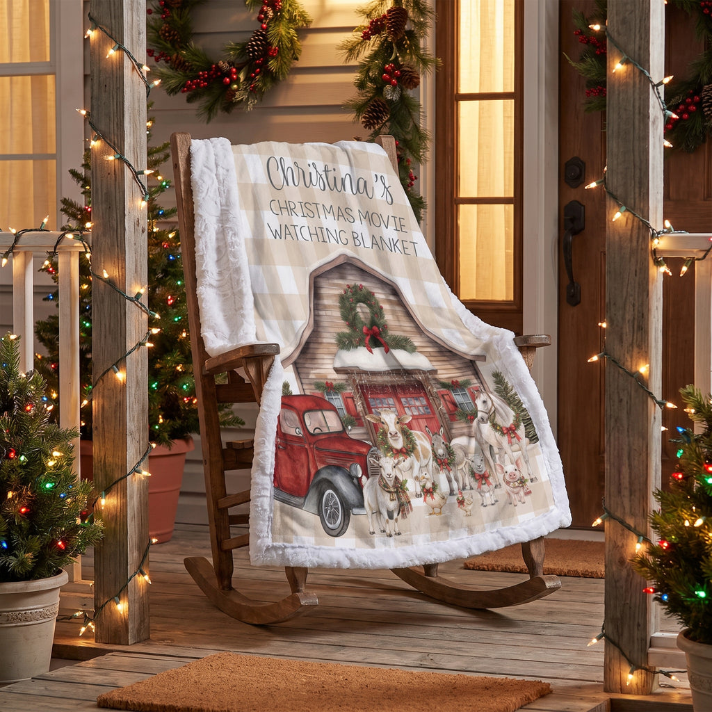 A personalized Christmas-themed fleece blanket draped over a wooden rocking chair on a porch. The blanket features a white and beige gingham border, the text 'Christmas Movie Watching Blanket' with a photo placeholder, and an illustration of a red truck, Santa, and holiday animals.