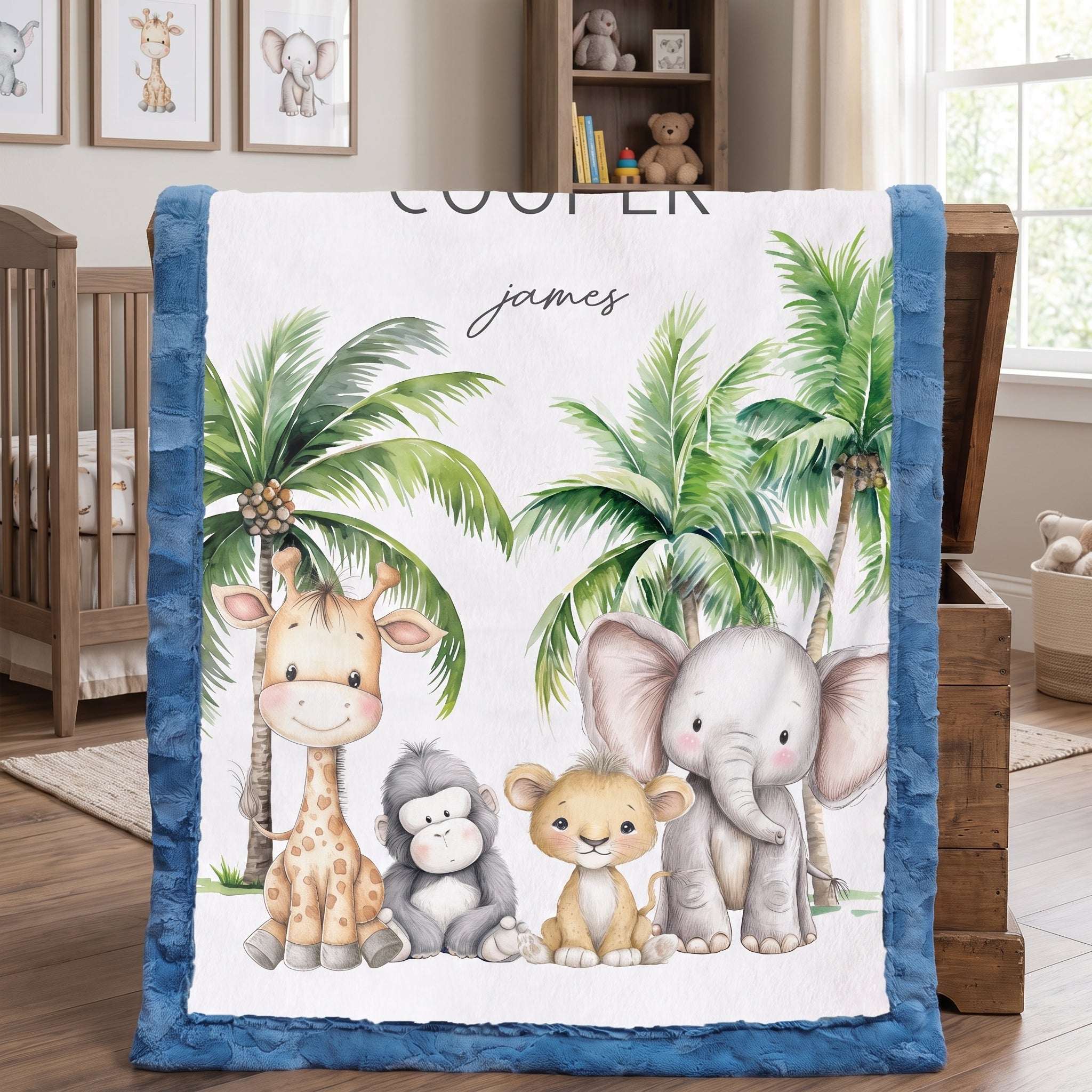 A personalized baby blanket with a blue minky border, featuring a white center printed with cartoon animals (giraffe, monkey, lion, elephant) and palm trees, with the name 'james' and the word 'COOLER' printed on it. The blanket is draped over a wooden chest in a nursery setting.