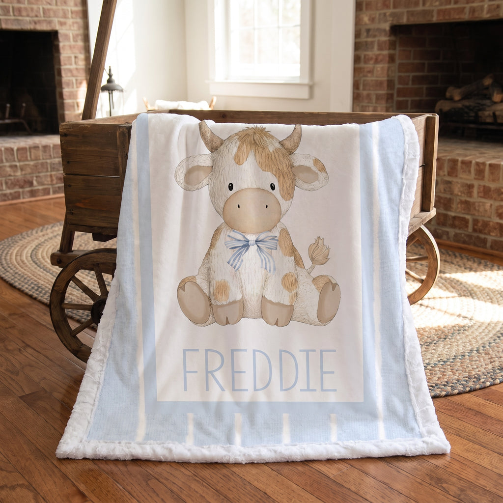 A personalized baby blanket featuring a cartoon cow named 'FREDDIE' in blue lettering, draped over a rustic wooden cart in a cozy, warm room with brick fireplaces and hardwood floors.
