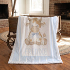 A personalized baby blanket featuring a cartoon cow named 'FREDDIE' in blue lettering, draped over a rustic wooden cart in a cozy, warm room with brick fireplaces and hardwood floors.