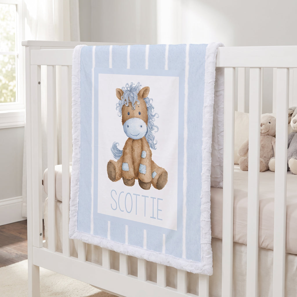 A personalized baby blanket draped over a white crib, featuring a watercolor illustration of a brown horse with a blue mane and the name 'SCOTTIE' printed below.