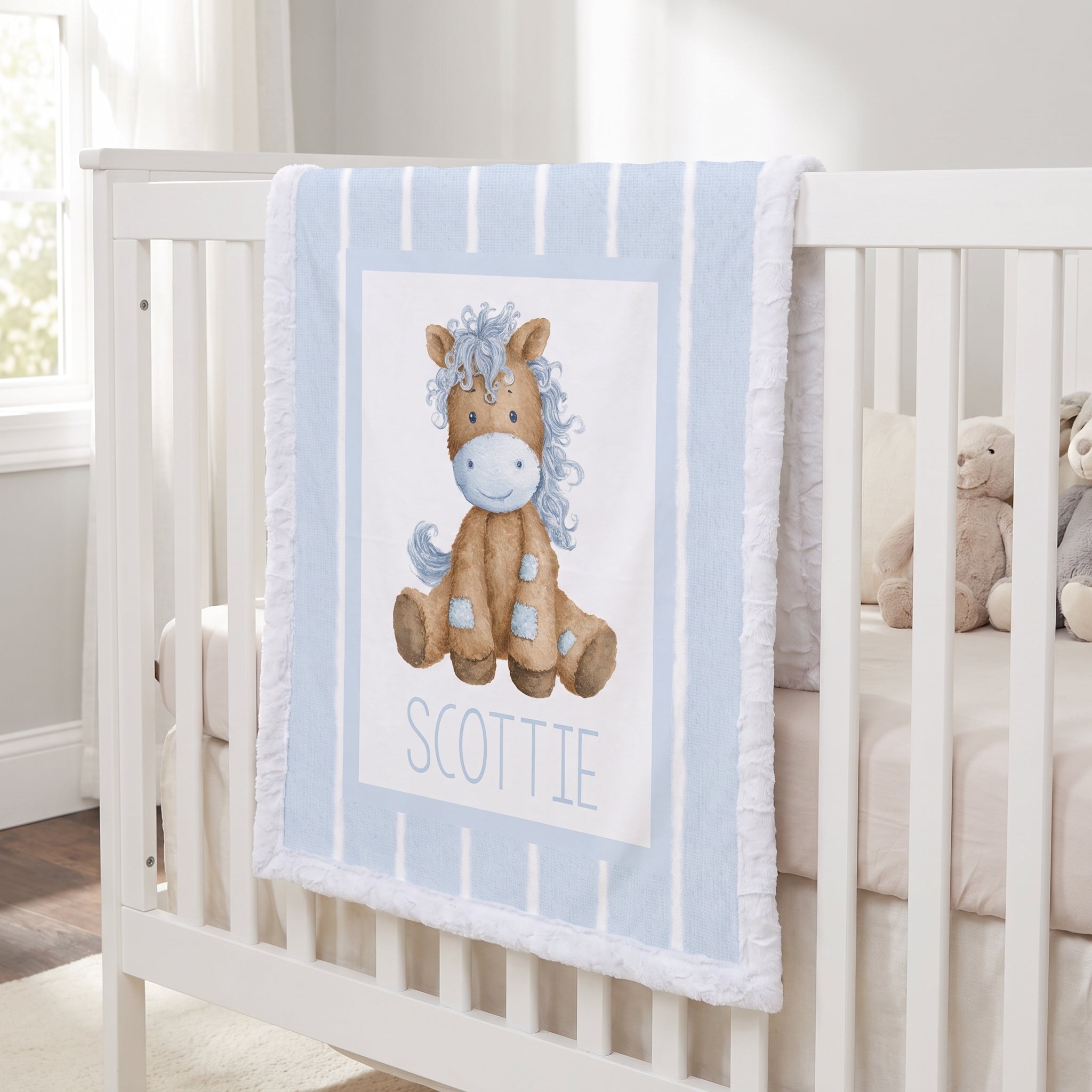 A personalized baby blanket draped over a white crib, featuring a watercolor illustration of a brown horse with a blue mane and the name 'SCOTTIE' printed below.