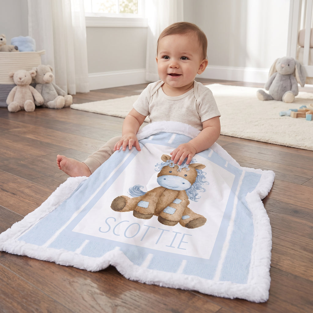 A baby sitting on a wooden floor in a nursery, wrapped in a personalized light blue and white blanket featuring a brown horse illustration and the name 'SCOTTIE'.