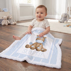 A baby sitting on a wooden floor in a nursery, wrapped in a personalized light blue and white blanket featuring a brown horse illustration and the name 'SCOTTIE'.