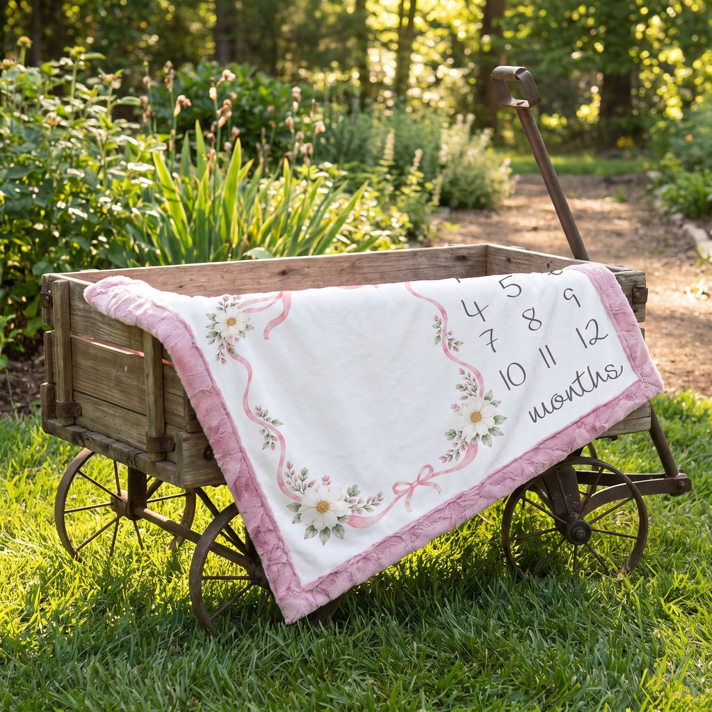 A white baby quilt with pink floral borders and a pink ruffled edge is draped over a rustic wooden wagon outdoors on a grassy lawn. The quilt has a printed black calendar from 1 to 12 months and the word 'months' written on it.
