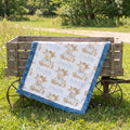 A baby quilt with a white background, blue ruffled border, and a repeating print of cartoon cows named Freddie, draped over a rustic wooden wagon in a grassy outdoor setting.