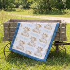 A baby quilt with a white background, blue ruffled border, and a repeating print of cartoon cows named Freddie, draped over a rustic wooden wagon in a grassy outdoor setting.