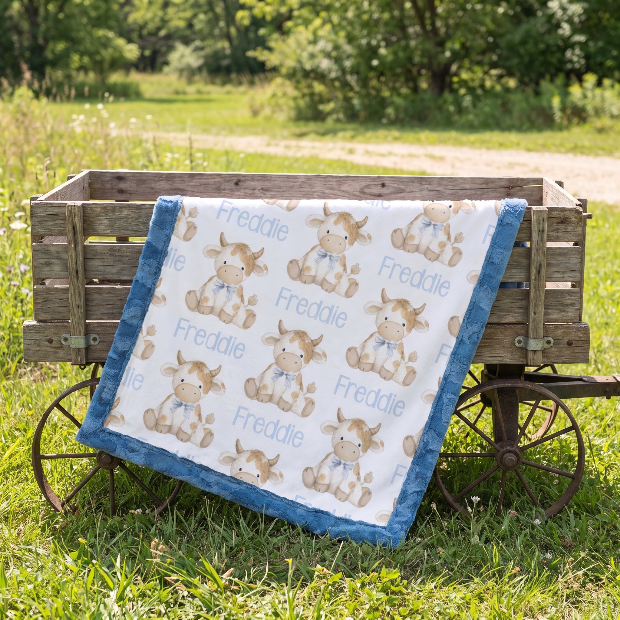 A baby quilt with a white background, blue ruffled border, and a repeating print of cartoon cows named Freddie, draped over a rustic wooden wagon in a grassy outdoor setting.