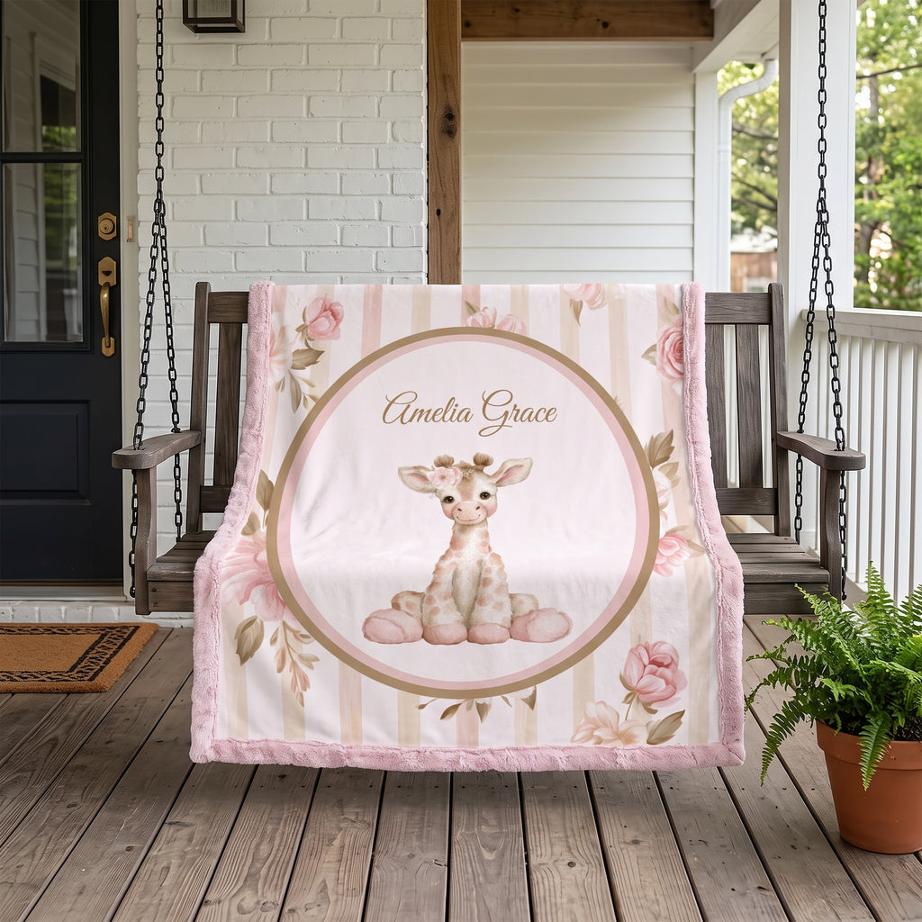 A personalized baby blanket draped over a wooden porch swing, featuring a cute cartoon giraffe in a floral crown inside a circular frame, pink roses, and the name 'Amelia Grace' printed at the top. The blanket has a soft pink faux-fur border and is set on a rustic wooden porch with a potted fern nearby.