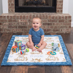 A baby sits on a personalized baby play mat in front of a brick fireplace. The play mat has a blue border and a white center printed with cartoon teddy bears, the name 'Freddie', and toys including a blue elephant, a colorful block toy, and a small ball.