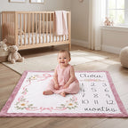 A baby in a pink dress sits on a personalized nursery floor mat that reads 'Clara 12 months' with floral designs and numbers, in a bright nursery with a wooden crib and teddy bears.